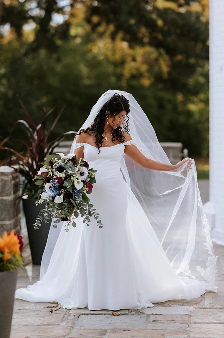 Bride in a strapless crepe wedding dress with off-shoulder lace sleeves, is holding her veil and a bouquet on a stone path.