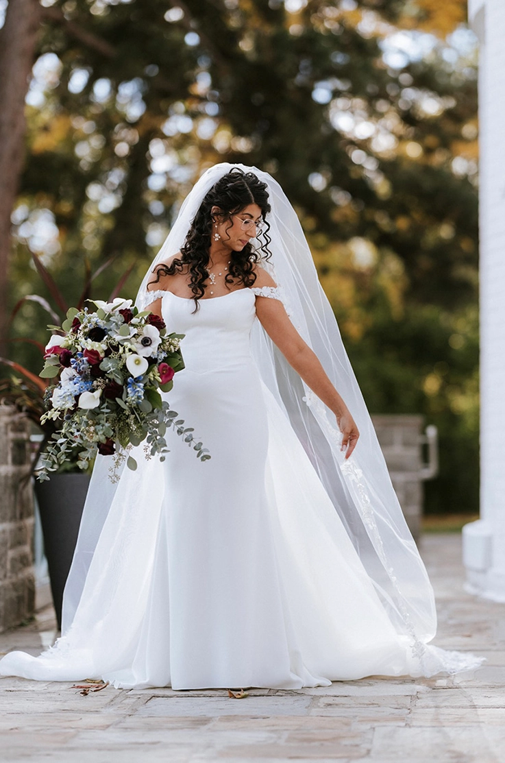 Bride poses in a scoop neck crepe gown with a fit and flare skirt, off-shoulder sleeves and a veil outside the venue.