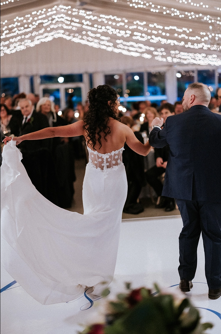 Bride in a crepe and 3D lace wedding gown, is holding up her train in a ballroom tent and dancing under string lights. 