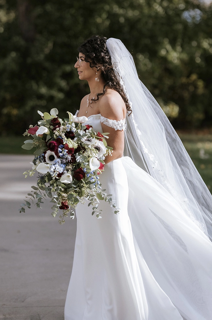 Smiling bride in a crepe fit and flare gown with lace sleeves and a veil, is holding a large bouquet outdoors in sunlight.