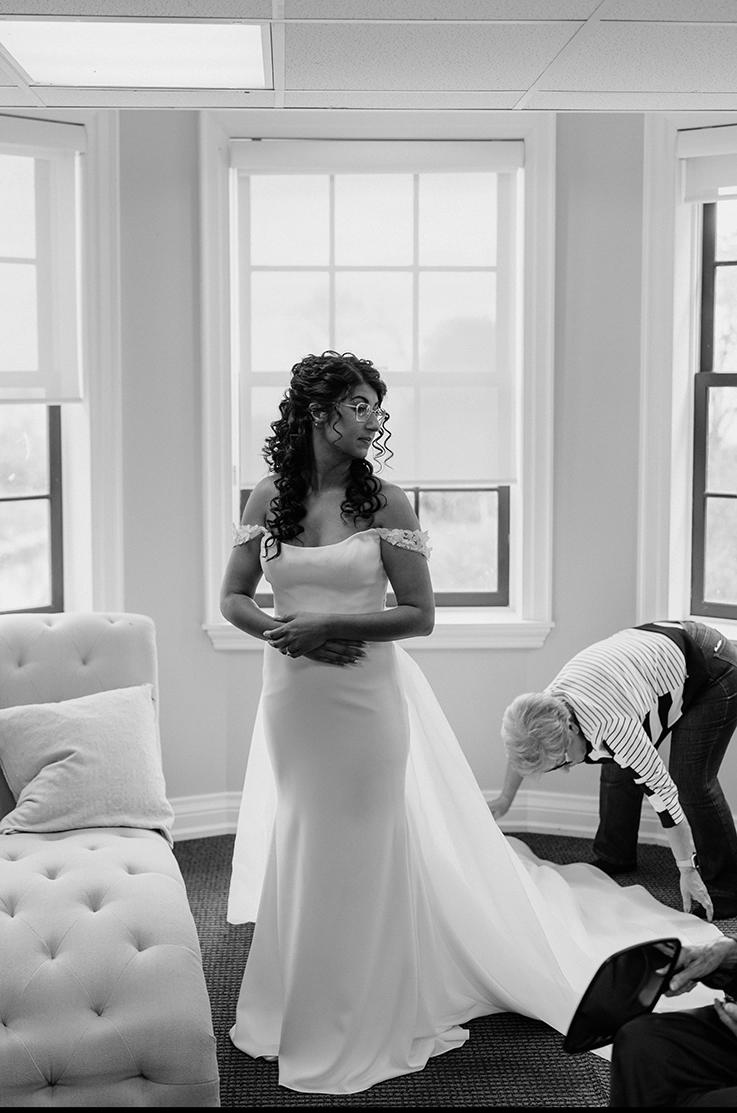Bride is getting ready in a crepe fitted gown with flower lace on the sleeves, standing by open windows in black and white. 