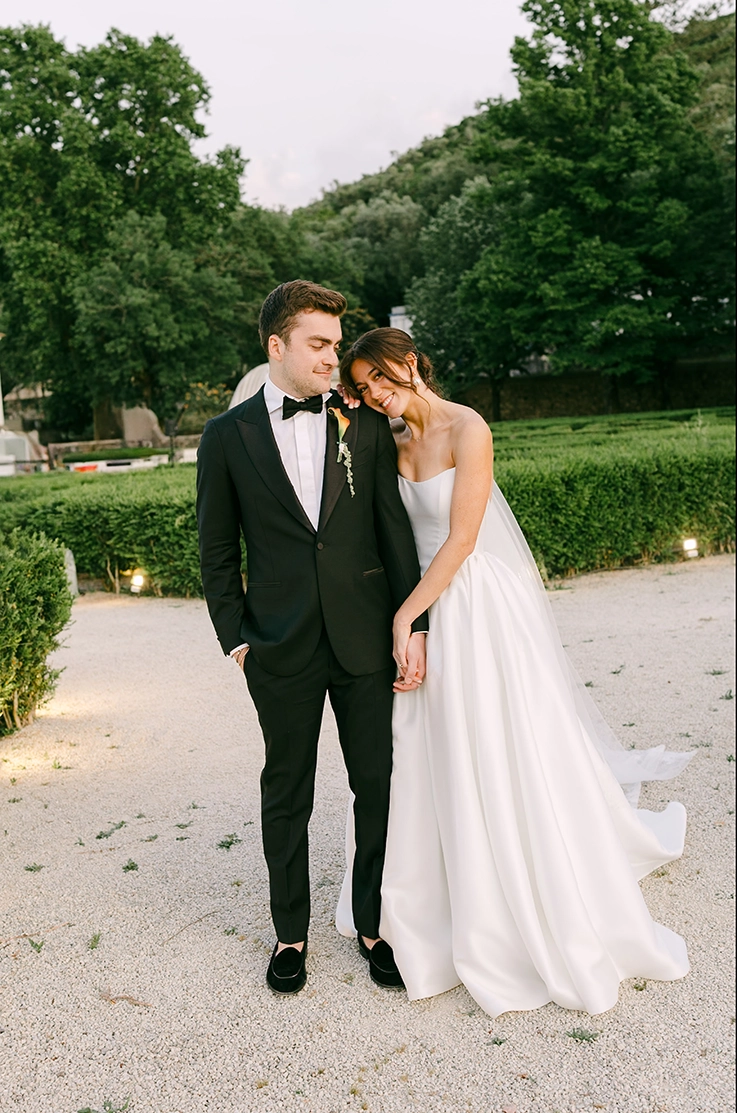 Bride smiles in a strapless mikado wedding dress with a gathered skirt and a veil, next to groom in manicured gardens.