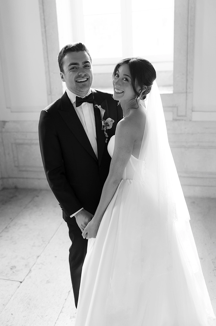 Groom poses with bride in a scoop neck mikado gown with a veil and a full gathered skirt, in a sunlit hall in black and white.