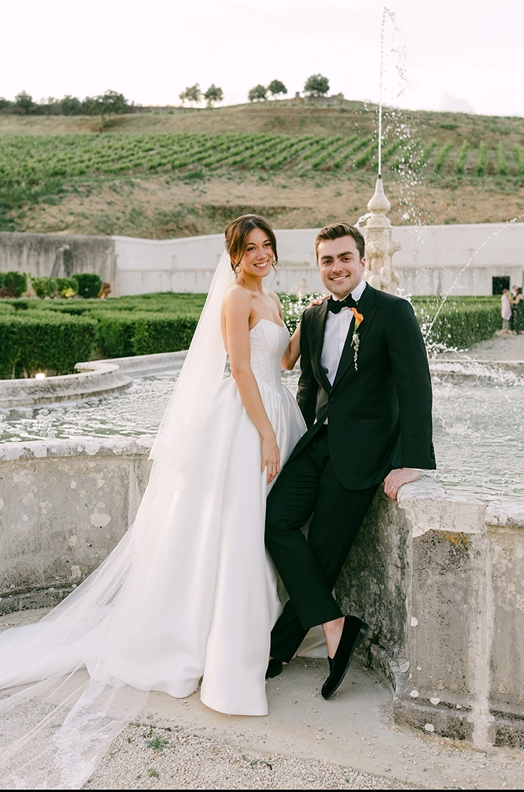 Bride in a mikado gown with princess seams on the bodice, a scoop neck and veil, stands by a fountain with her groom. 