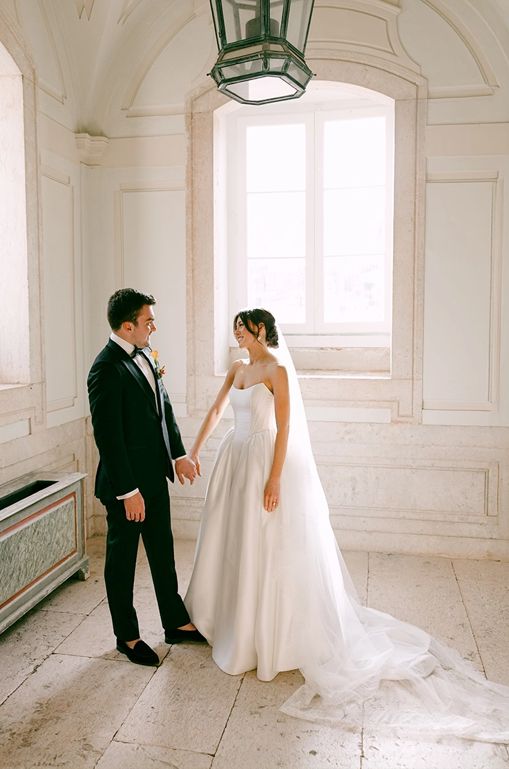 Bride in a mikado ball gown with a full skirt, princess seams and a veil, faces her groom in front of bright windows. 
