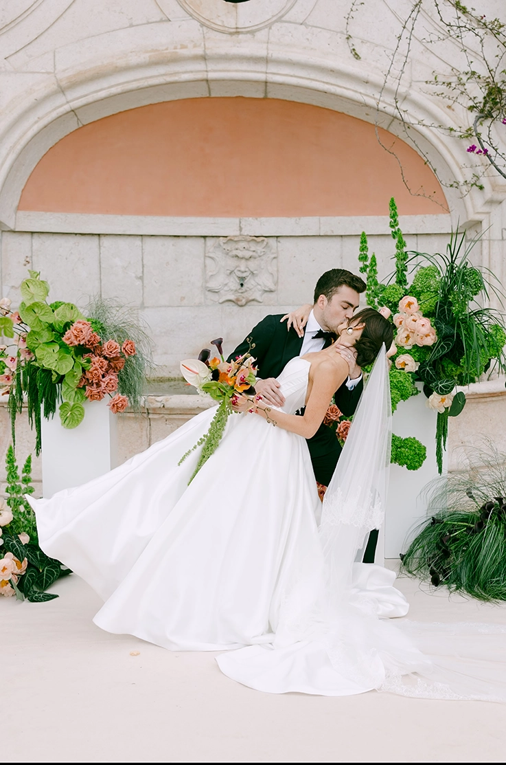 Groom dips his bride in a mikado gown with princess seams, a full skirt and a veil, at a stone fountain decorated with flowers.