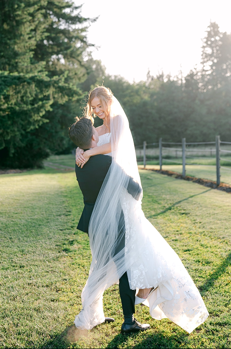 Groom picks up his bride in an embroidered tulle gown with a flowing veil, in a sunny open field by a wood fence.