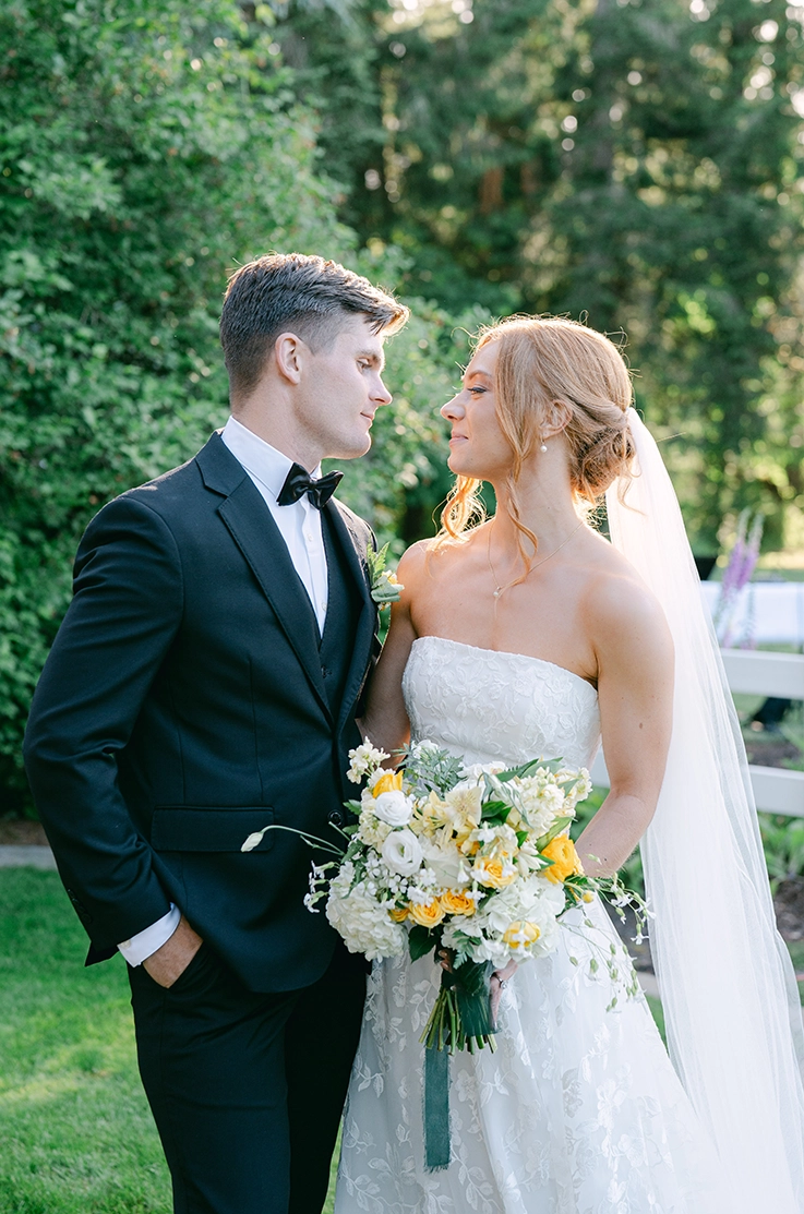 Bride in a strapless embroidered tulle and lace gown with a veil, holds a bouquet and faces her groom outdoors in sunlight.