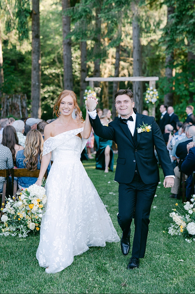 Bride in a strapless floral lace wedding dress with embroidered tulle, walks hand in hand with groom in the aisle in a forest.