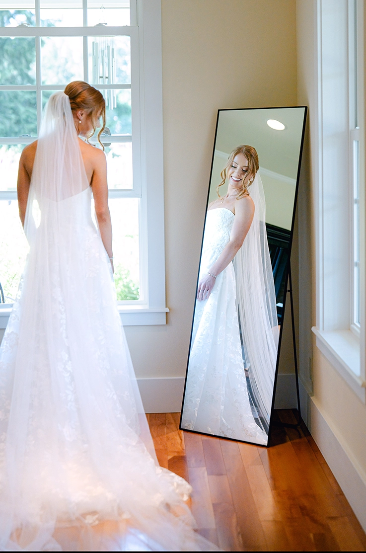  Bride looks at her reflection in a mirror, wearing a floral lace A-line gown with a long veil, in front of open windows.