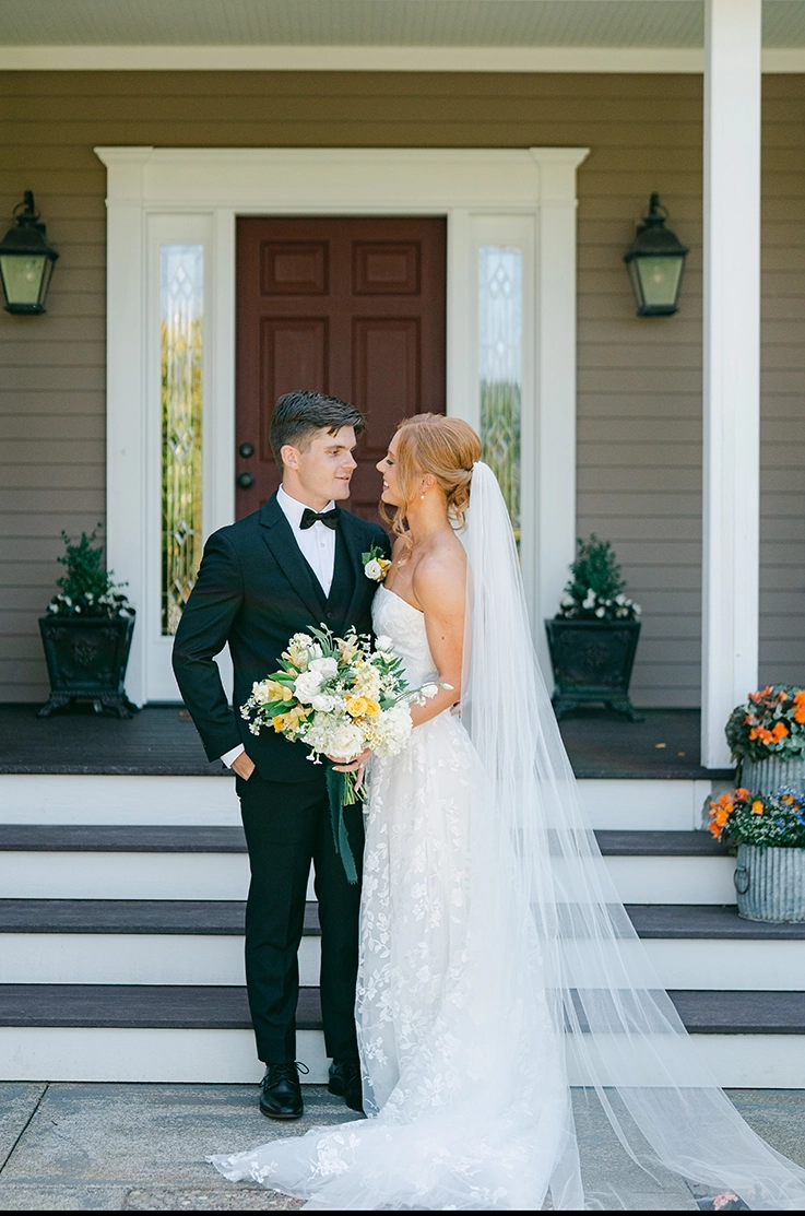 Bride in a strapless floral lace and tulle gown with a long veil, holds a bouquet at the bottom of steps with her groom.