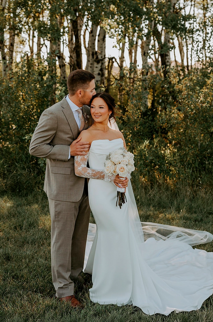 Bride in a fit and flare satin wedding dress with a veil and lace sleeves holds a bouquet, and poses with groom in a forest. 