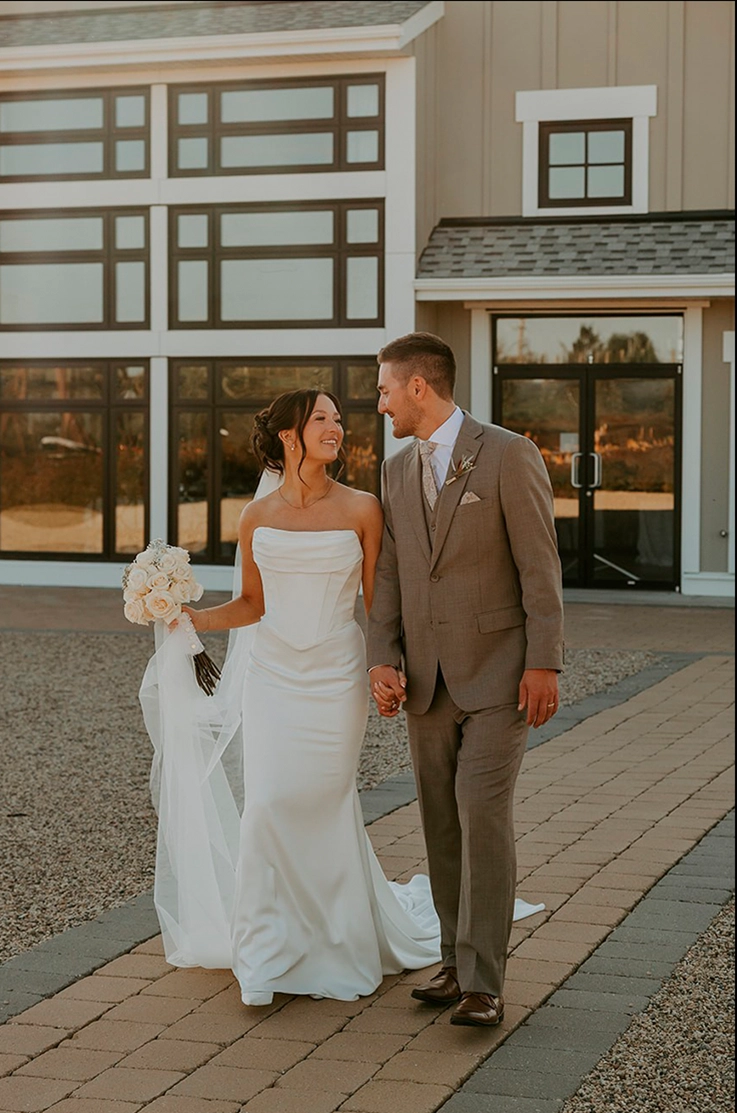 Bride in a satin draped neck gown with a fit and flare skirt and veil, walks with her groom smiling as they exit the venue. 