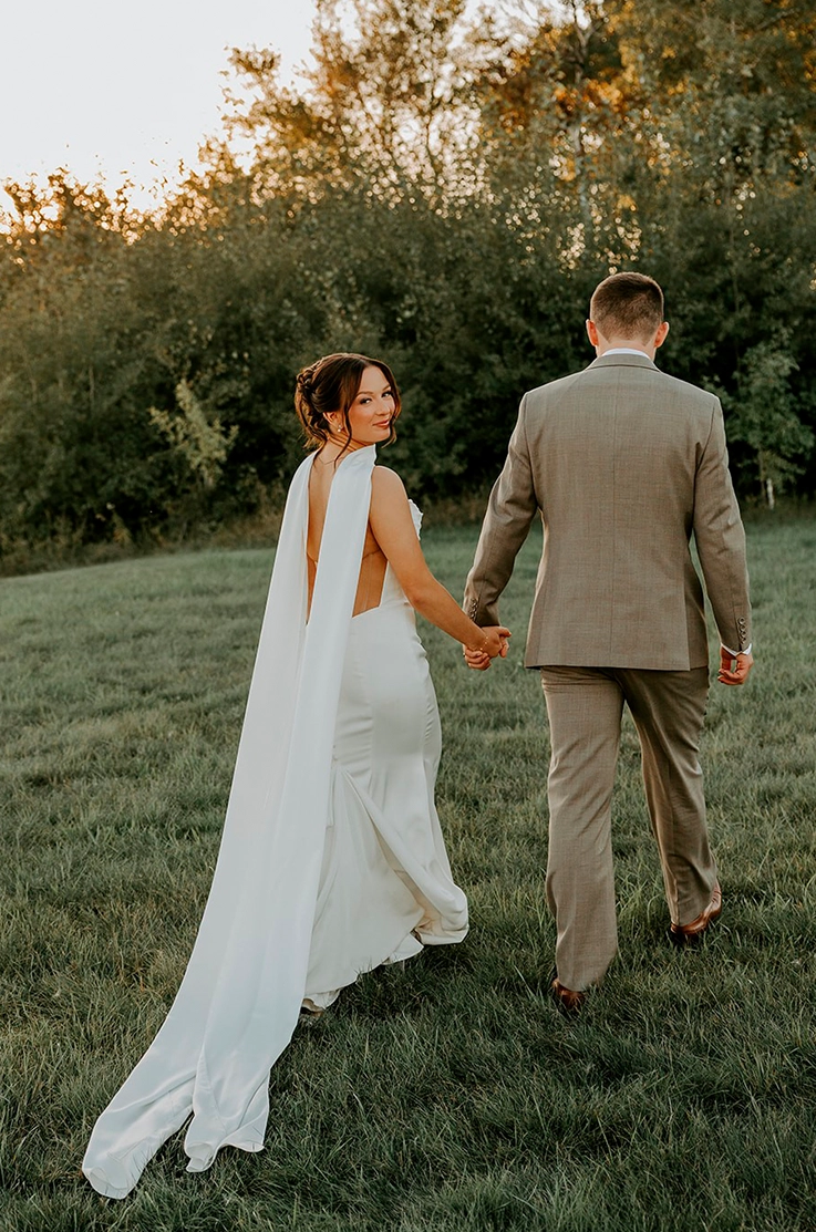 Bride looks back in a satin fit and flare gown with nude back detail and a scarf, walking hand in hand with her groom in a field.