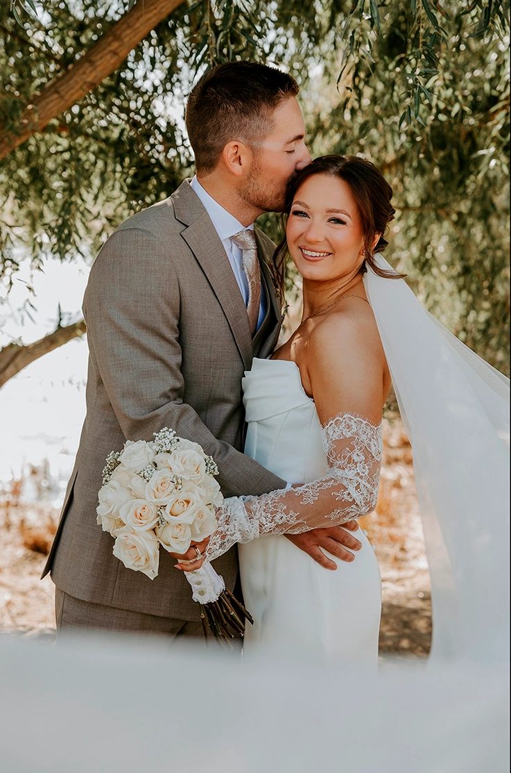Groom embraces bride in a strapless fitted gown with a draped neckline, sleeves and a veil, holding a bouquet under willow trees.