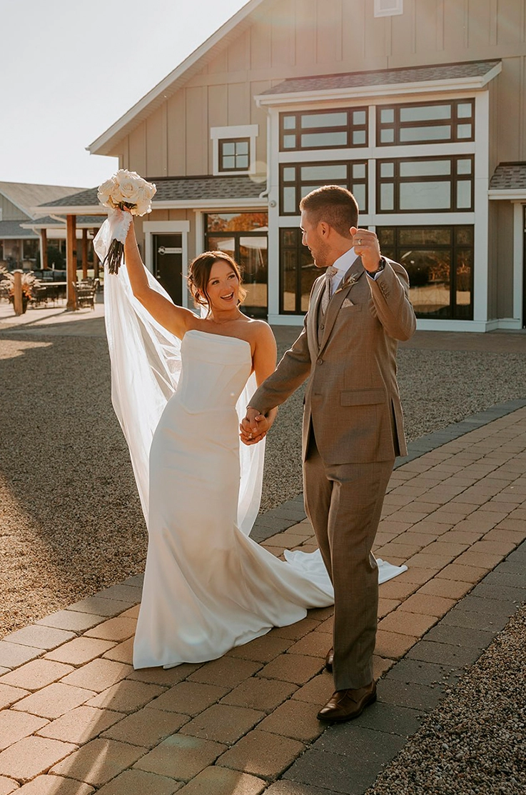 Bride celebrates in a scoop draped neck gown with a flowing veil and fitted skirt, alongside her groom outside the venue