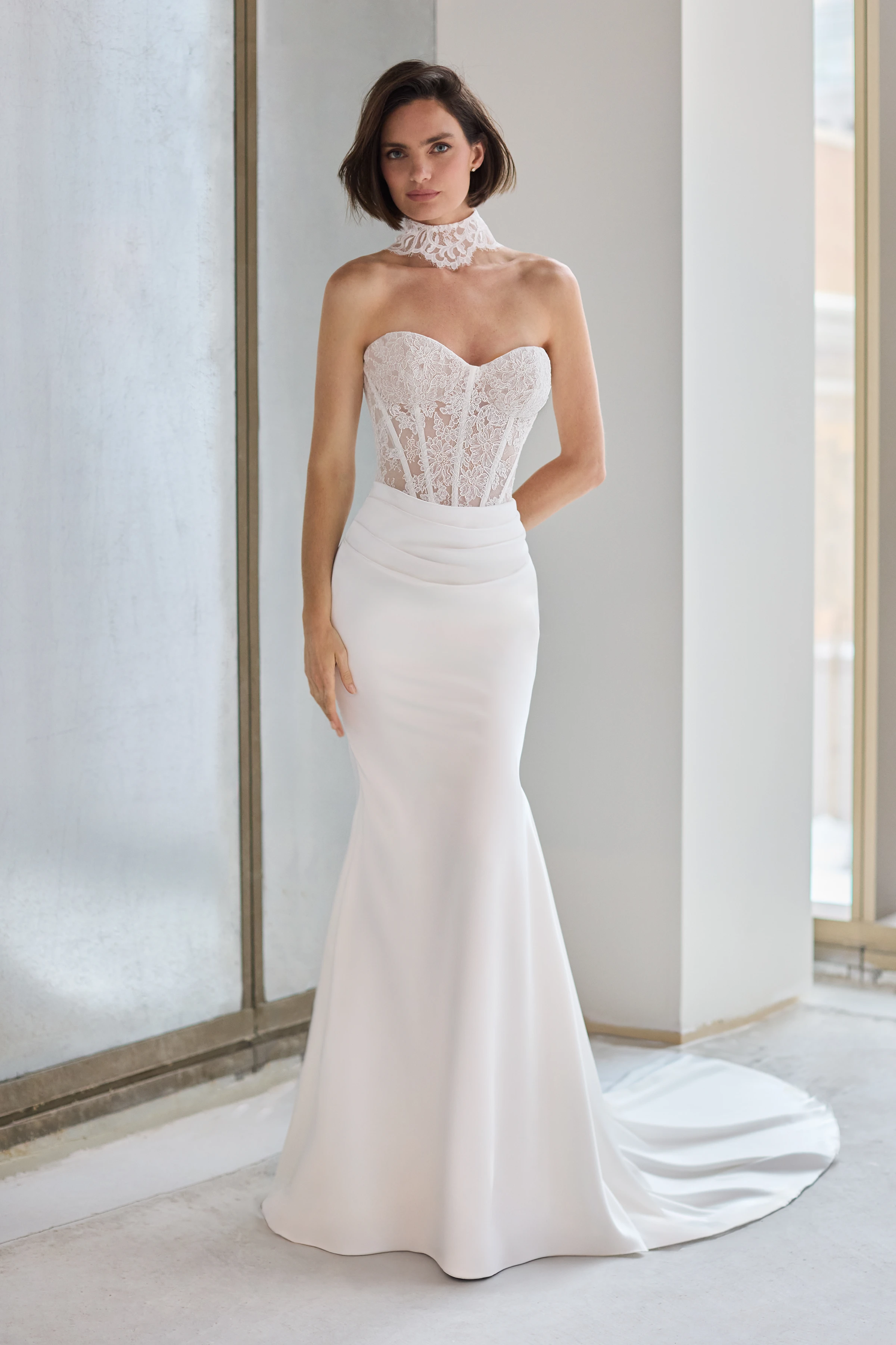 Front view of a strapless wedding dress with a sheer Lace bodice, a draped fit and flare Charmeuse skirt, and a Lace choker, in a studio.