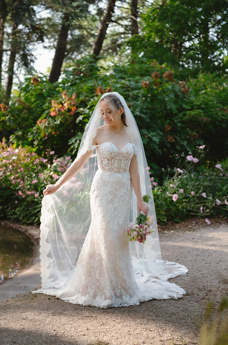  Bride in a strapless lace wedding dress with draped sleeves, poses holding her veil and a bouquet on a garden path. 