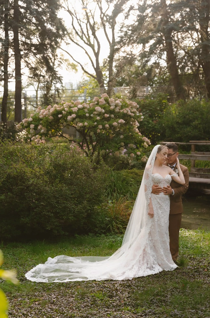Groom holds bride in an embrace in a garden, wearing a fitted lace gown with a sweetheart neckline and a long lace-trim veil. 
