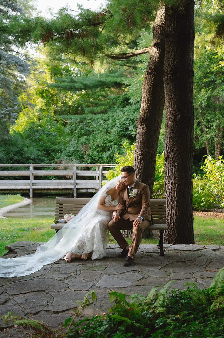 Bride in a strapless lace gown with a flowing veil, is posing with the groom as they sit on a bench beneath tall pine trees. 