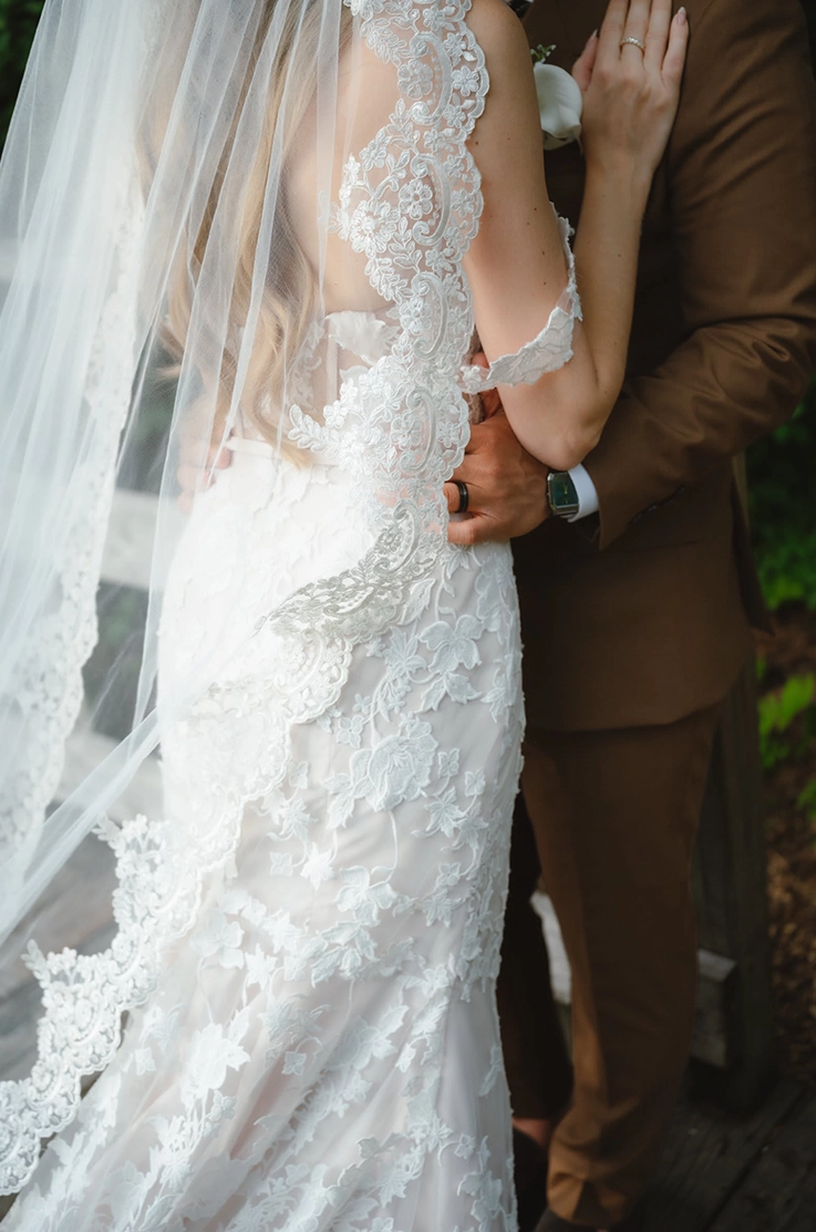 Close-up of bride in a low back lace gown with draped sleeves, a veil and fit and flare skirt, facing her groom. 