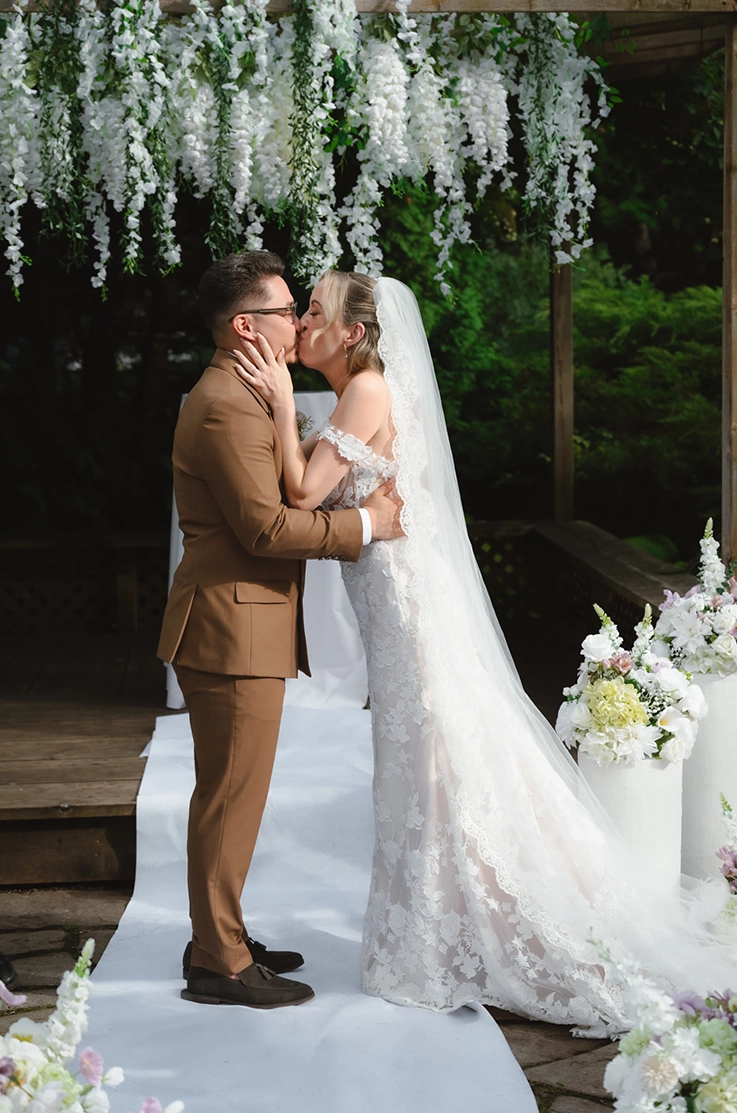 Bride in a lace fit and flare gown with draped sleeves and a long veil, kisses her groom at altar, decorated with white flowers.