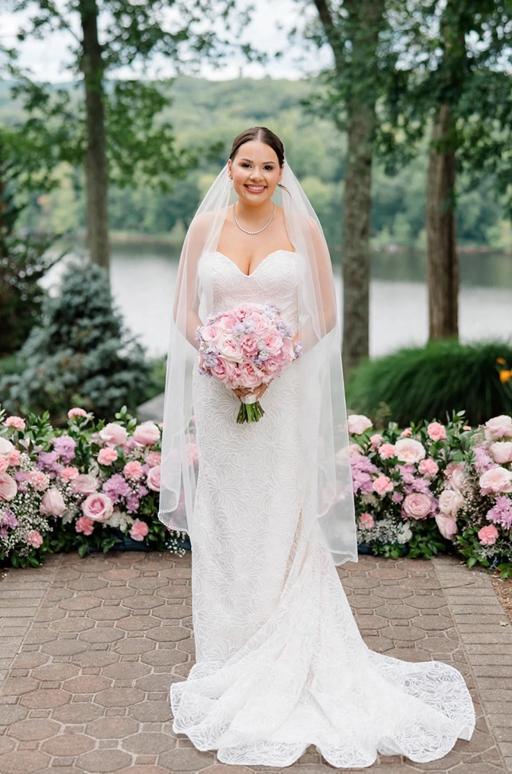 Bride smiles in a strapless corded tulle wedding dress with a veil and holds a bouquet, surrounded by pink florals outdoors.
