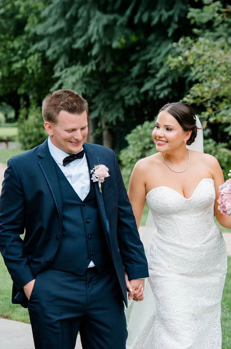 Bride in a strapless corded tulle gown with a veil and sweetheart neckline, walks hand in hand with groom on a garden path.