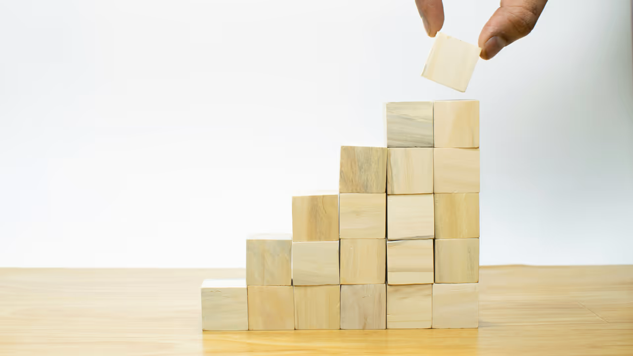 Wooden square blocks forming steps with a hand placing the final block on top.