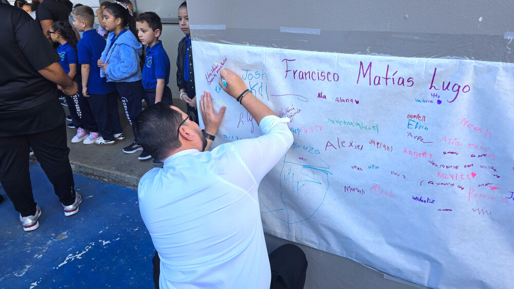 Subsecretario del Departamento de la Vivienda firma un cartel conmemorativo de la Escuela Elemental Francisco Matías Lugo mientras estudiantes en uniformes azules observan durante el recibimiento del primer día de clases en Carolina, Puerto Rico.