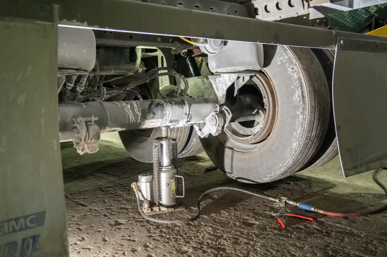 Axle lifted by bottle jack under semi trailer, “IMC” on mudflap; tools and air hose staged for wheel-end service.