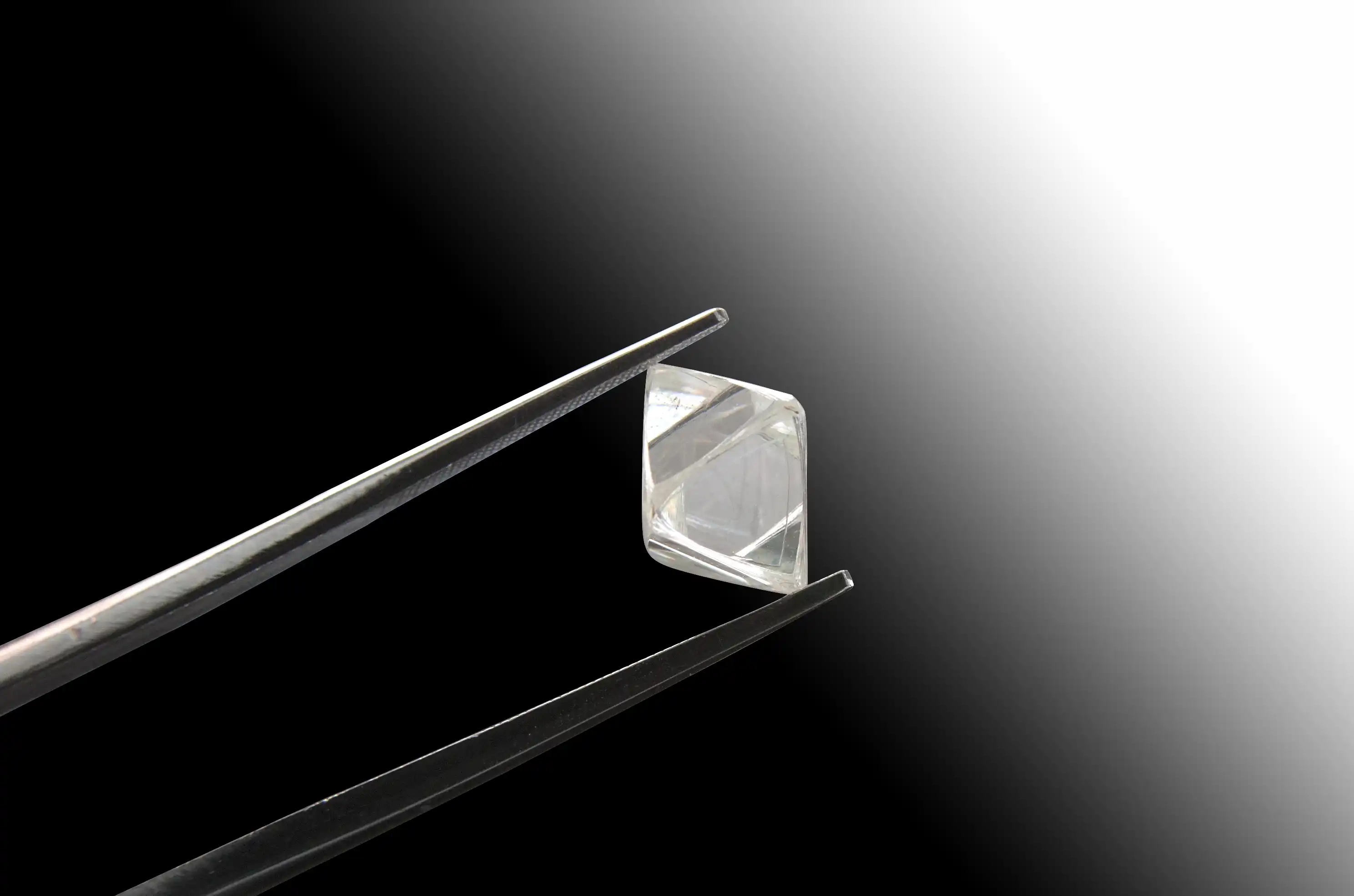 Kleine, transparente, facettierte Kristallspitze, die mit einer Pinzette vor einem schwarz-weißen Hintergrund gehalten wird.