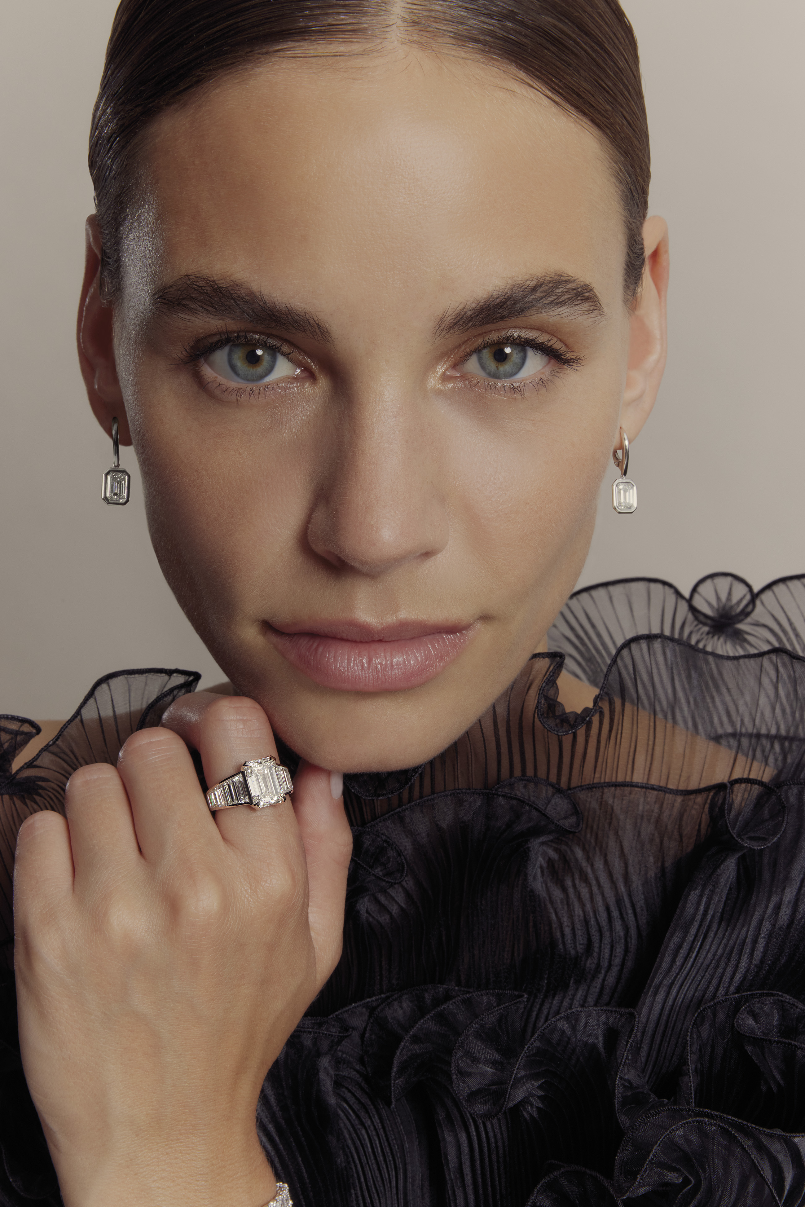 Nahaufnahme einer Frau mit grün-blauen Augen, die silbernen Diamant-Ohrringe und einen großen Diamantring an der Hand trägt, bekleidet mit einem schwarzen, transparenten, rüschigen Oberteil.