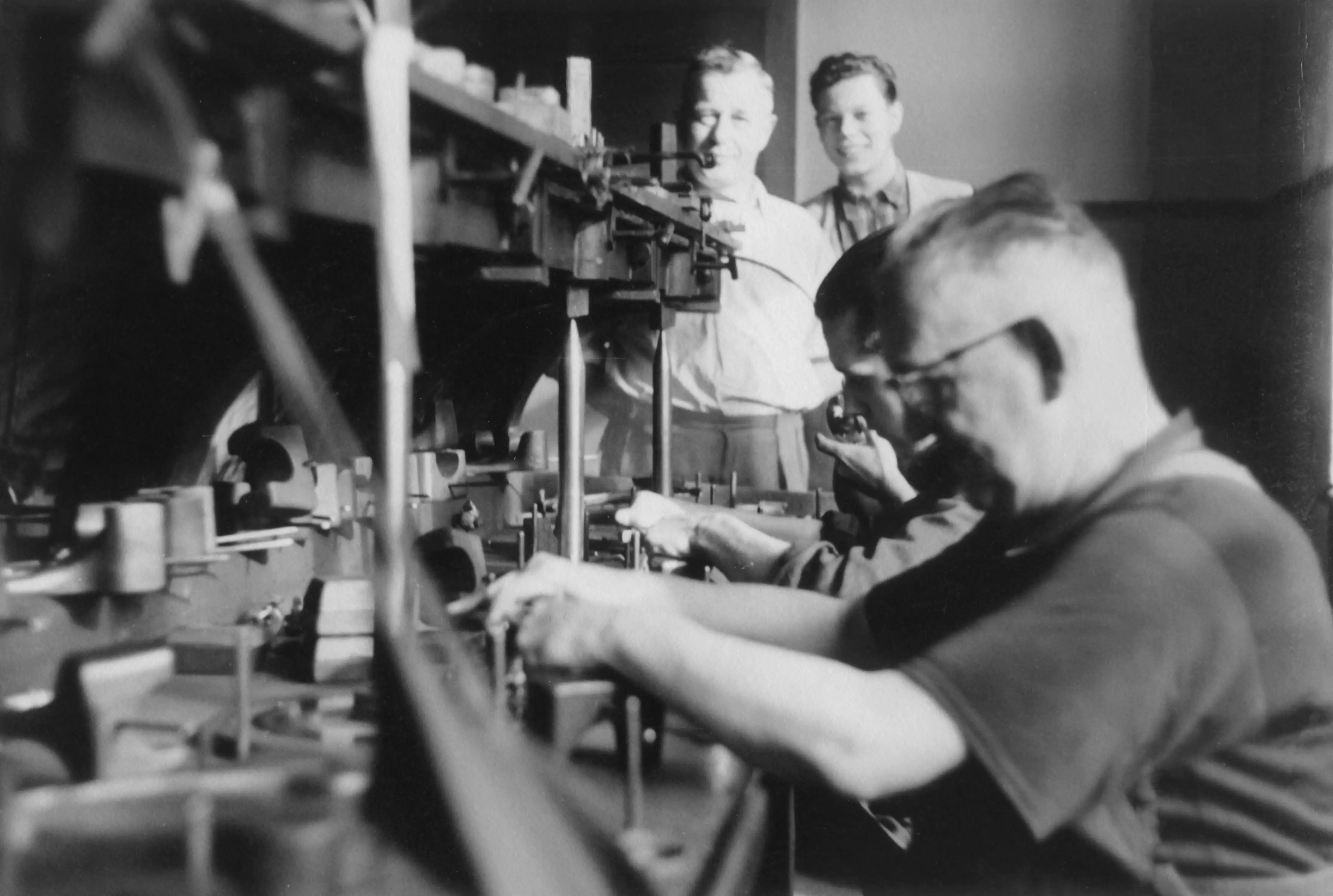 Schwarzweißfoto von vier Männern an einer Werkbank, zwei arbeiten konzentriert, zwei beobachten und lächeln im Hintergrund.