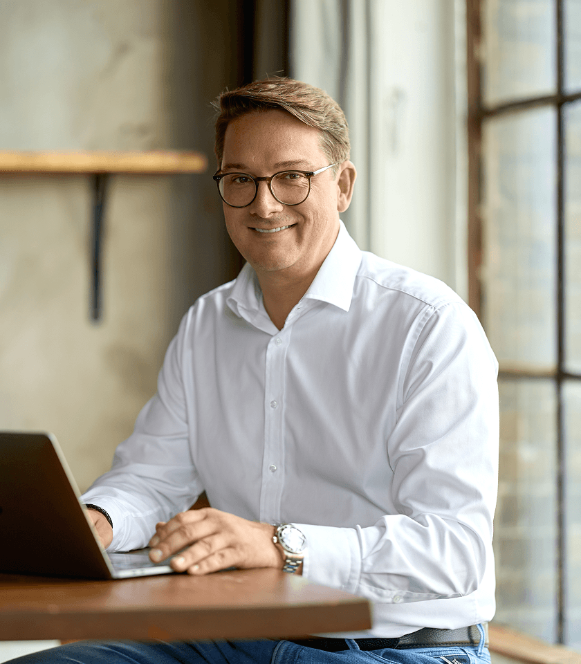 Man wearing glasses and a white shirt smiling while using a laptop at a wooden table near a window.