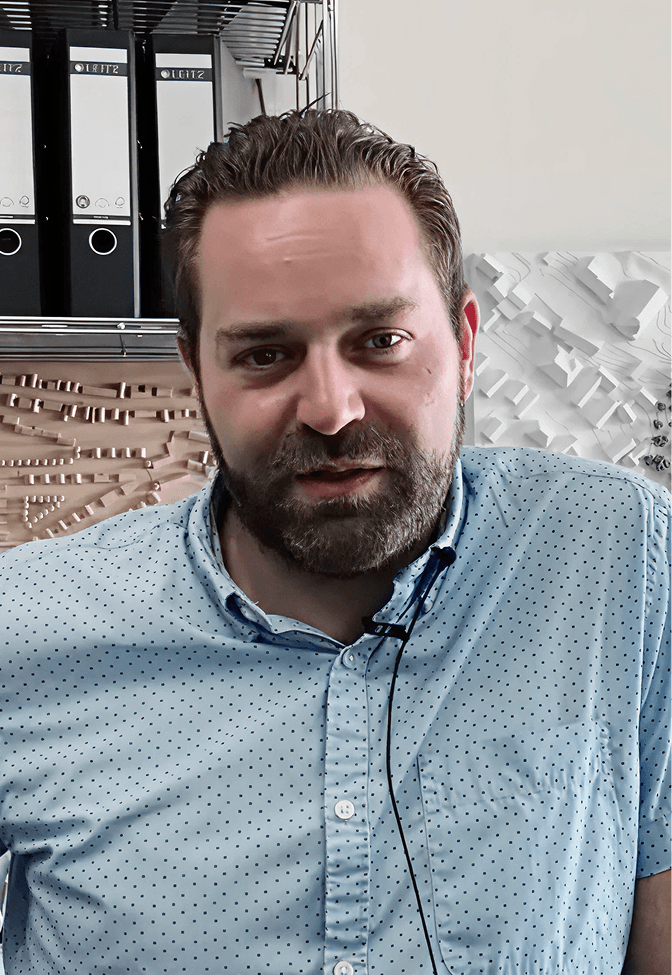 Man with beard and short hair wearing a light blue polka dot shirt in an office setting with binders and architectural models behind him.