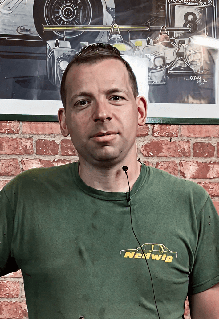 Man wearing a green shirt with text 'Norwig' and a microphone headset standing in front of a brick wall and a framed racing car poster.