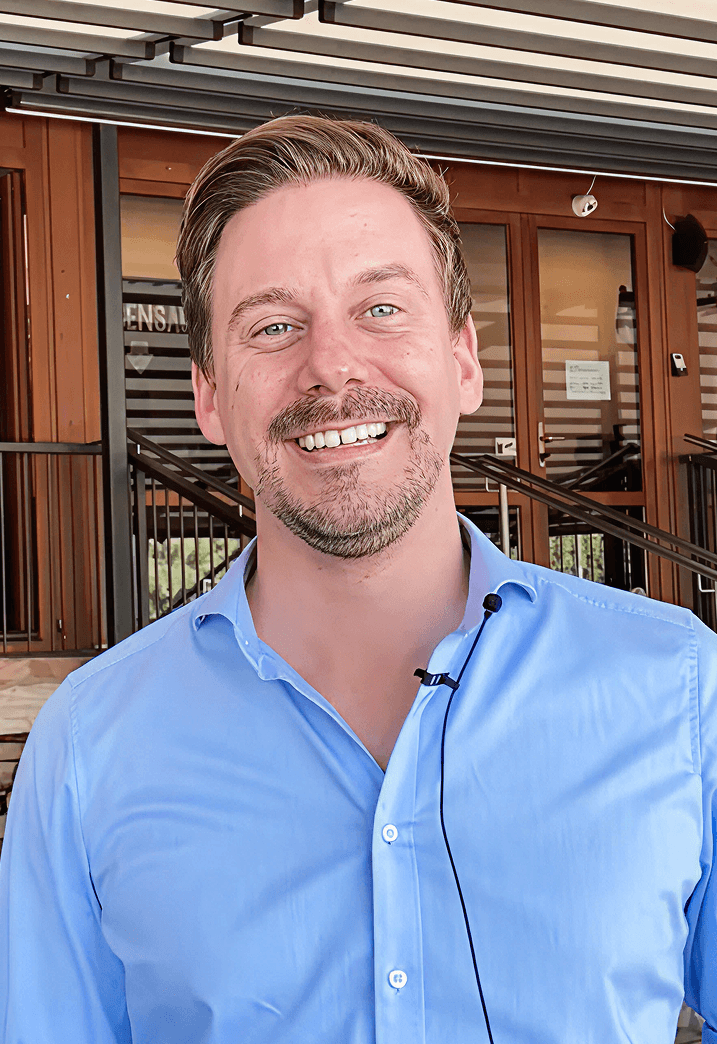 Smiling man with light brown hair and beard wearing a light blue button-up shirt with a microphone clipped on, standing in front of a building entrance with glass doors and metal railings.