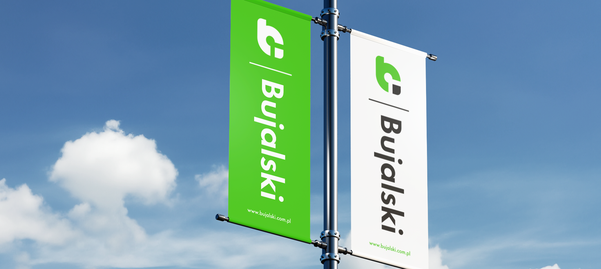 Two vertical flags on a pole against a blue sky, one green and one white, both displaying the Bujalski logo and website.