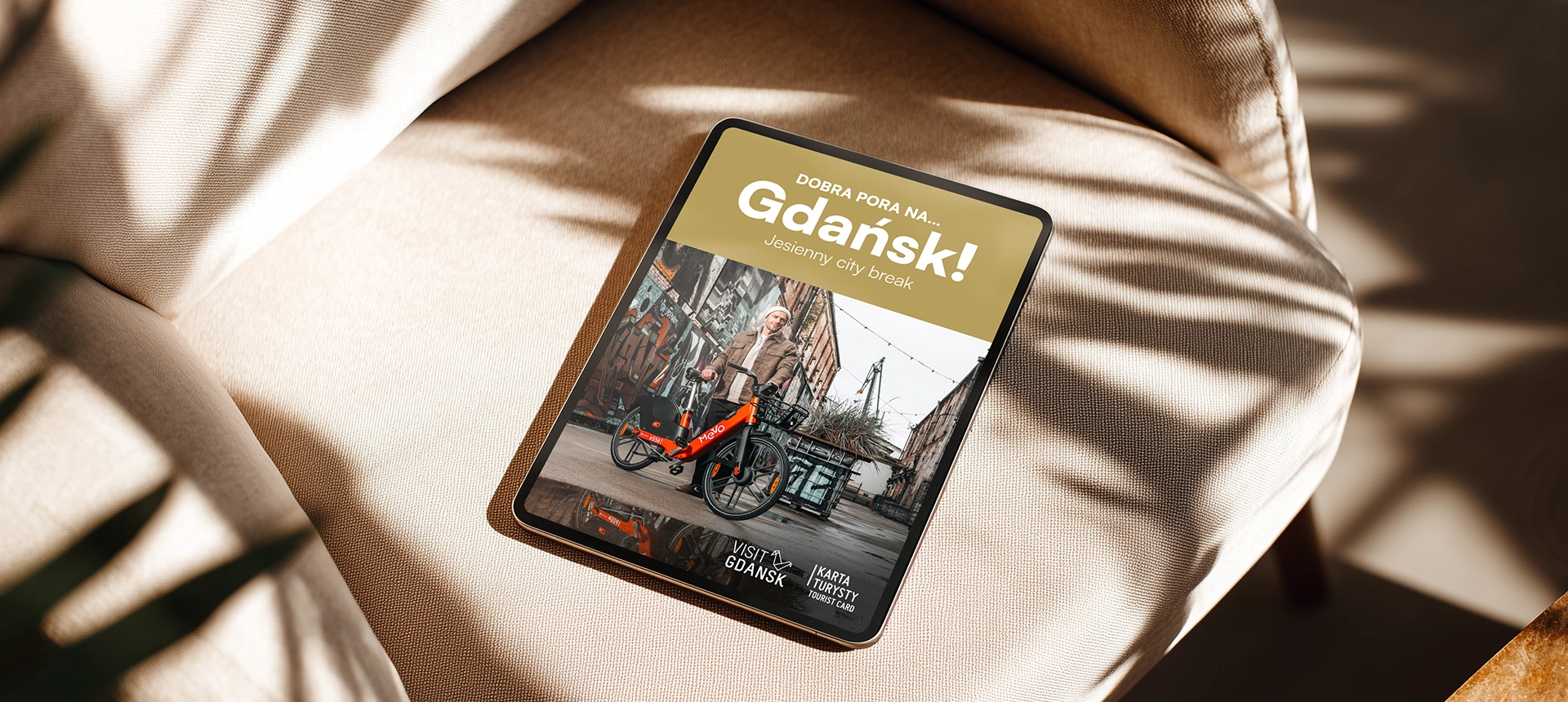 Tablet on beige fabric chair showing a promotional image for Gdańsk with a man posing next to an orange Mevo bike in an urban setting.