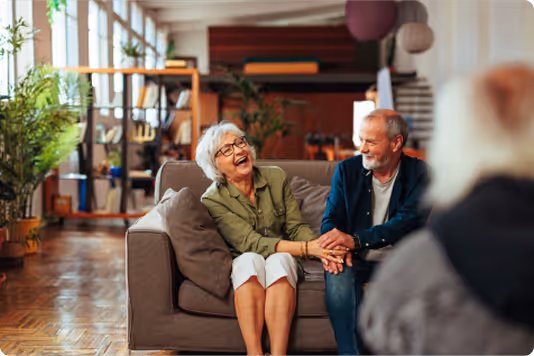 Elderly couple sitting on a sofa in a cozy living room, laughing and holding hands.