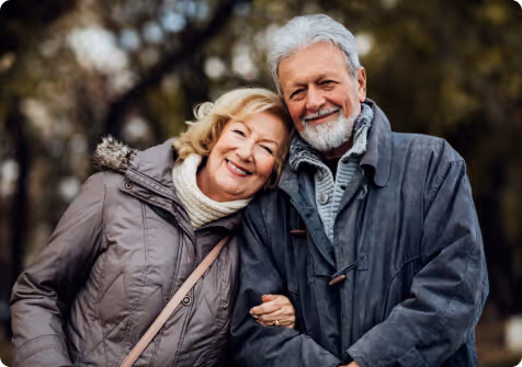 Smiling elderly couple in warm coats embracing outdoors with trees in the background.
