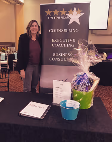 Woman standing next to a display table with a banner reading 'Five Star Relationships' offering counselling, executive coaching, and business consulting, along with a gift basket and informational materials.