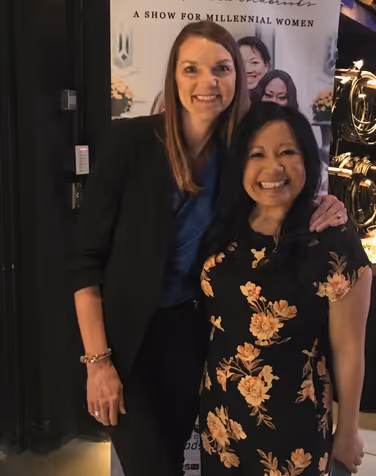 Two smiling women posing together indoors, one wearing a black blazer with a blue top, the other in a black floral dress.