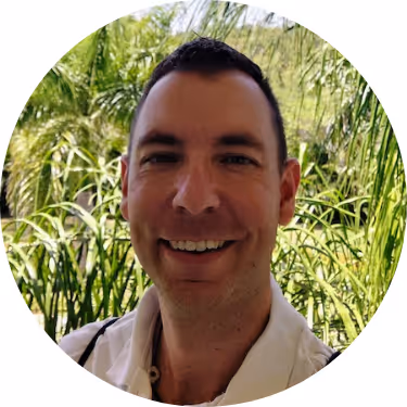 Smiling man with short dark hair in a white shirt standing outdoors with green plants in the background.