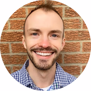 Smiling man with short hair and beard wearing a blue and white checkered shirt in front of a brick wall.