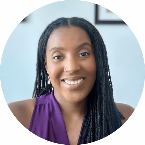 Smiling woman with long black dreadlocks wearing a sleeveless purple top against a light blue background.