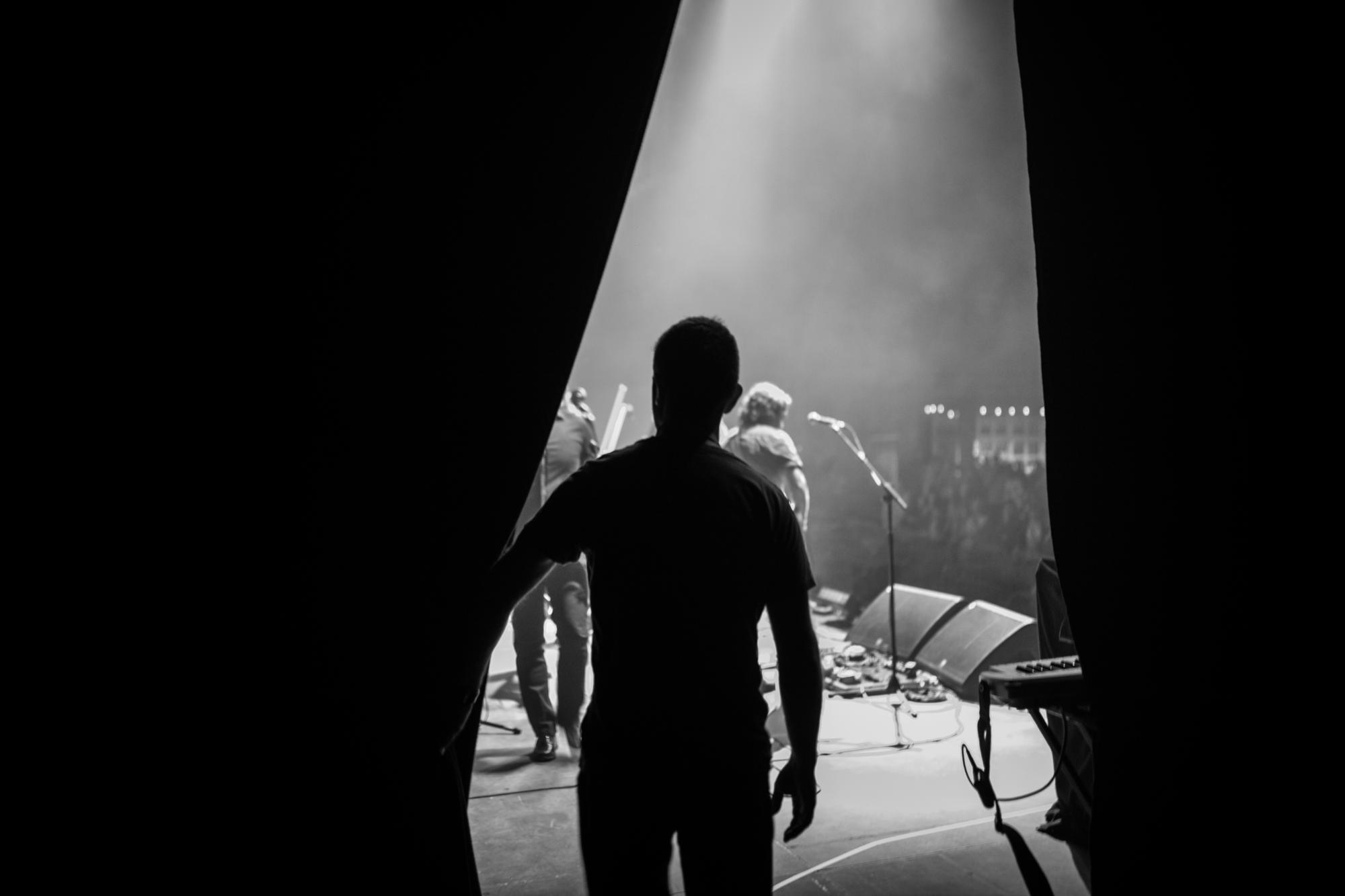 Silhouette of a person standing behind stage curtains watching musicians perform on a foggy concert stage.