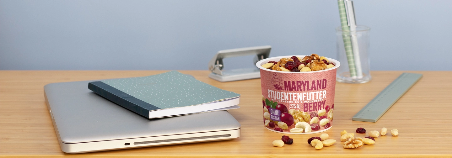 Snack-Becher mit Maryland Studentenfutter und verstreuten Nüssen auf einem Schreibtisch neben Laptop, Notizbuch, Lineal und Stiftglas.