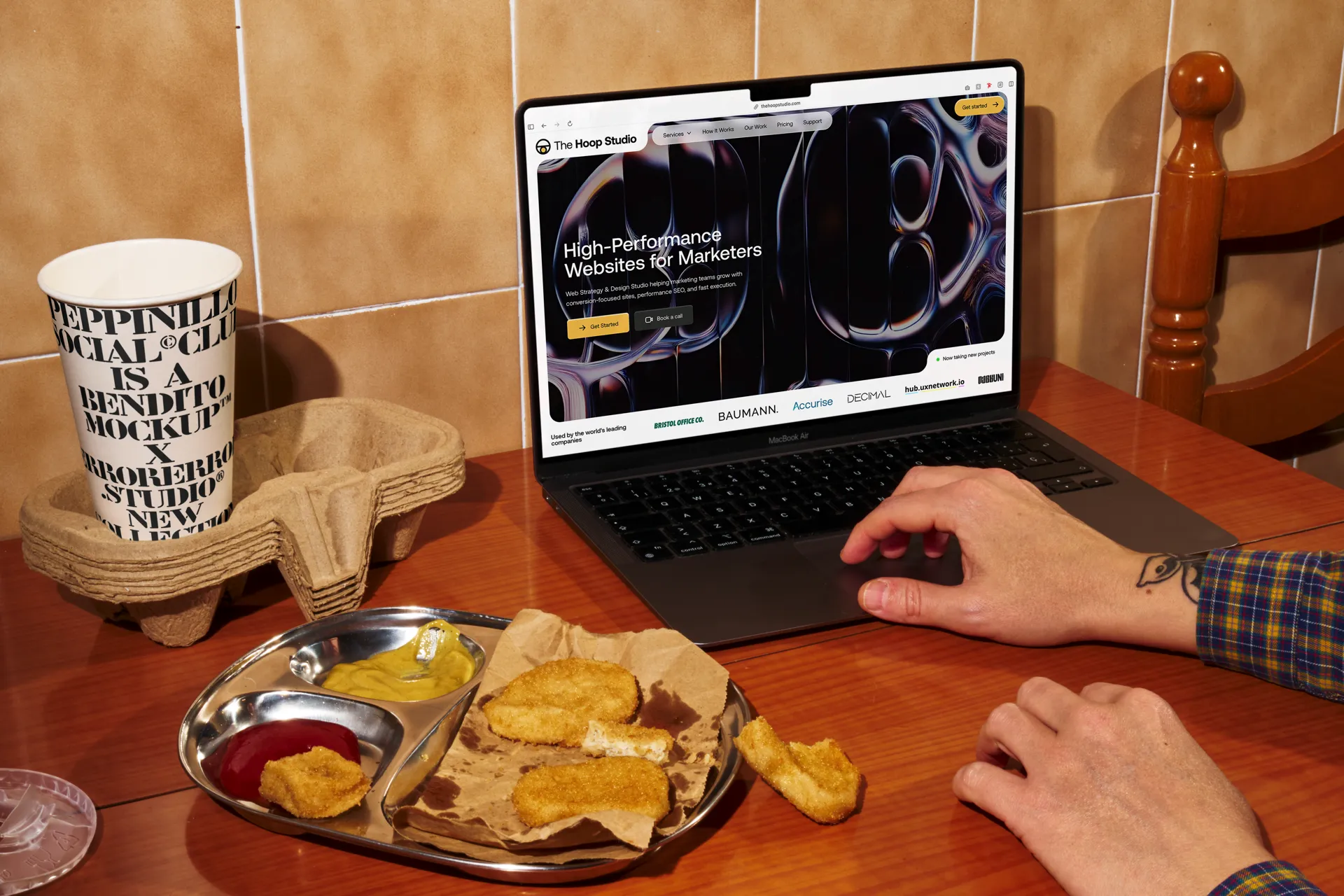 Person with tattooed wrist using a MacBook Air on a wooden table next to a tray with chicken nuggets, mustard, ketchup, and a paper cup in a holder.